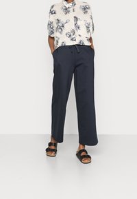 Chemise légère à manches courtes avec un motif floral en noir et blanc. Pantalons amples bleu marine foncé avec une taille à cordon. Sandales noires.