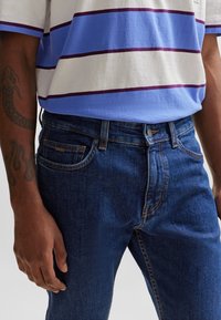 Denimjeans med en mörkblå tvätt, som har kontrastfärgade oranga sömmar, en framficka och en klassisk knappstängning. Matchas med en randig blå och vit t-shirt.