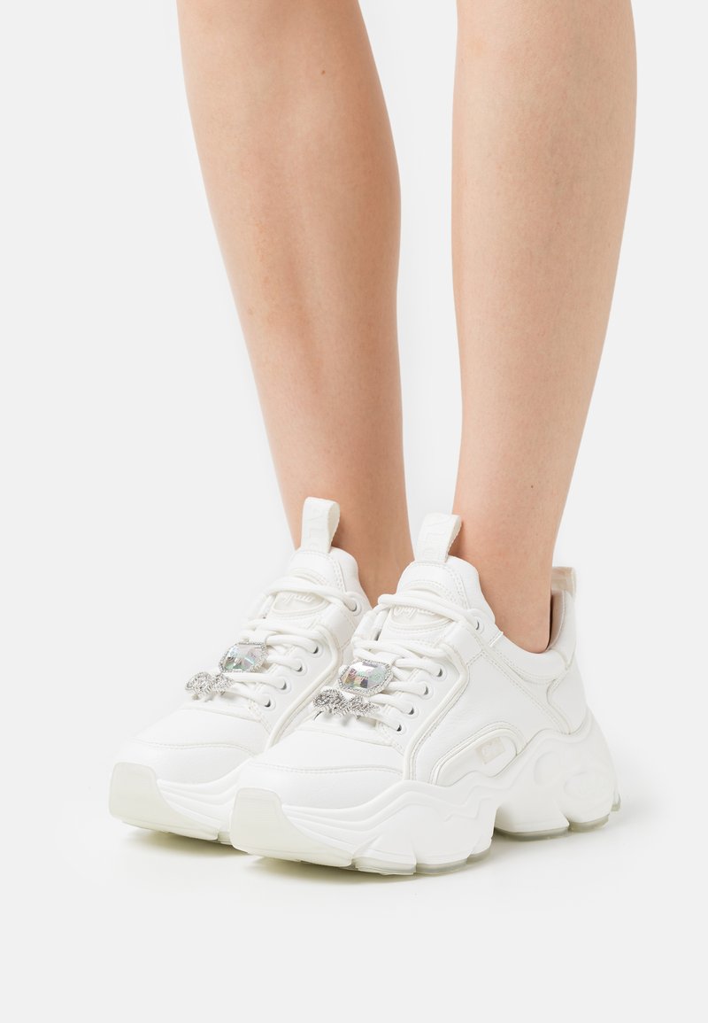 White sneakers made of smooth synthetic material with a chunky sole, featuring decorative crystal accents on the laces and pull tabs.