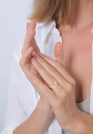 Gold ring with a wave design, featuring a small pearl accent. The model has well-groomed hands, showing natural nail polish.