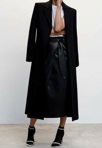 Manteau long noir à coupe ajustée, associé à une jupe en cuir noire avec un détail de taille noué. Bottines noires avec accents blancs.
