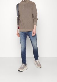 Person, der einen braunen langärmligen Reißverschluss-Pullover, blaue Jeans, beigen Sneaker trägt und eine kleine beigefarbene Umhängetasche mit marineblauem Riemen trägt.