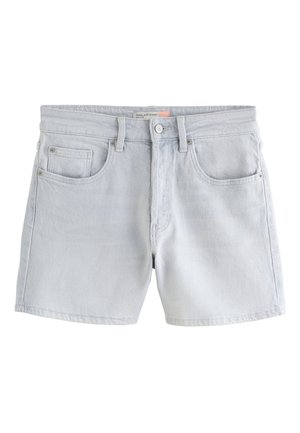 Vrouw met donker haar die een gele gehaakte trui met banaanpatronen en lichtgrijze korte broek draagt, staand met haar rug tegen een stenen muur.