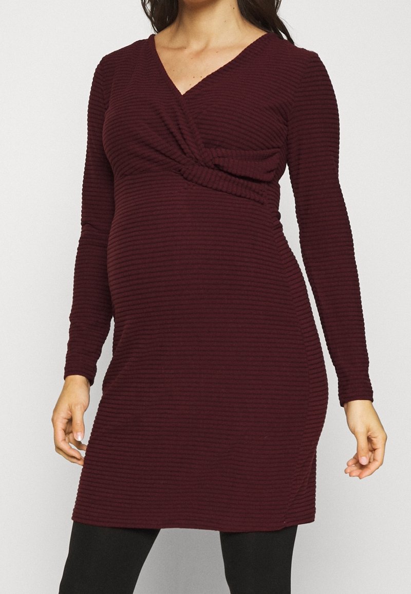 Femme portant une robe de grossesse à manches longues, côtelée, de couleur bordeaux foncé, avec un décolleté en V et un détail de tissu superposé sur la poitrine.