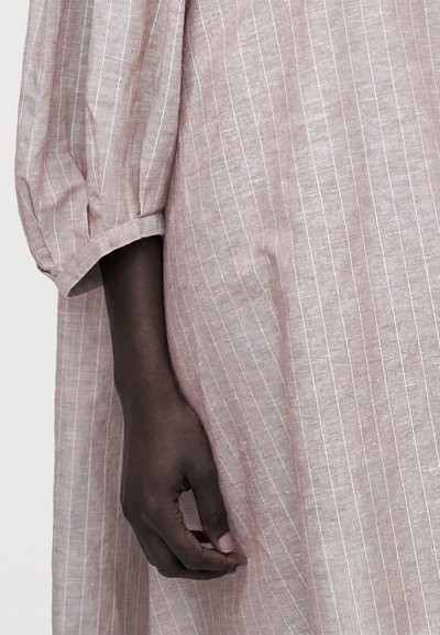 Χέρι με σκούρο δέρμα που ακουμπά σε ανοιχτό ροζ, ριγωτό, χαλαρό ύφασμα με σιρίτι, τονίζοντας την υφή και την αντίθεση στο ύφασμα και τους τόνους του δέρματος.