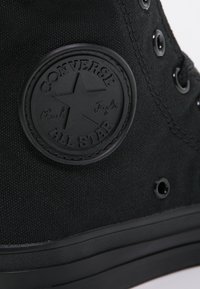 Schwarzer Canvas-High-Top-Sneaker mit einer strukturierten Stoffoberfläche, rundem Gummilogo-Patch, schwarzen Ösen und einer glatten Gummisohle.