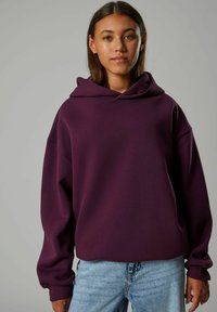 Lila übergroße Kapuzenjacke mit großer Fronttasche, lockerer Passform und gerippten Bündchen, kombiniert mit hellblauen Jeans. Glatte Textur.