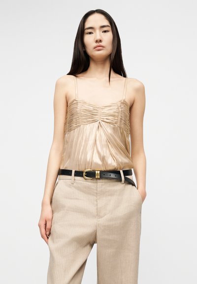 Νεαρή γυναίκα με μακριά σκουρόχρωμα μαλλιά, φορώντας μπεζ πλισέ σατινέ καμιζόλ, μπεζ παντελόνι και μαύρη ζώνη με χρυσή αγκράφα.
