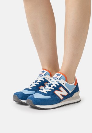 Par de zapatillas New Balance 574 en azul, azul claro, blanco y naranja, usadas en los pies sobre un fondo gris claro liso.