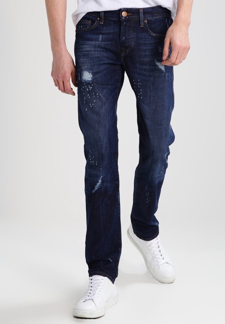 Jeans en denim bleu foncé à coupe slim, présentant des détails usés et une texture délavée, associés à des baskets blanches.