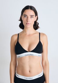 Donna con capelli corti e scuri che indossa un reggiseno nero di Calvin Klein e delle mutande abbinate, in piedi contro uno sfondo chiaro e semplice, rivolta in avanti.