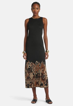 Žena koja stoji, nosi bezrukavu crnu midi haljinu s donjim dijelom u smeđem uzorku, crne sandale, zlatnu narukvicu, ogrlicu i naušnice.