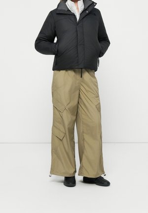 Personne portant une veste matelassée noire, un pantalon cargo beige à jambes larges avec des poches, et des chaussures noires, debout les mains dans les poches de la veste.