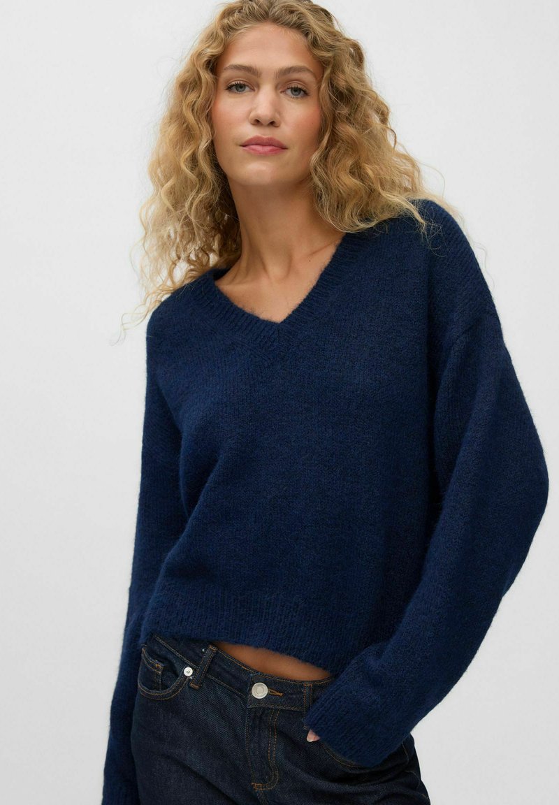 Femme blonde aux cheveux bouclés portant un pull bleu foncé à col en V et un jean foncé sur un fond uni.