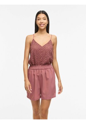 Młoda kobieta uśmiecha się, nosi bezrękawnik w kolorze mauve z wzorem i dopasowane szorty z wysokim stanem na jednolitym białym tle.