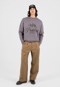 Grauer gestrickter Pullover mit dem Text "Verboten" und einem Olivenbaumdesign; kombiniert mit beigefarbenen, hochgeschnittenen, gefältelten Hosen und schwarzen Schuhen.