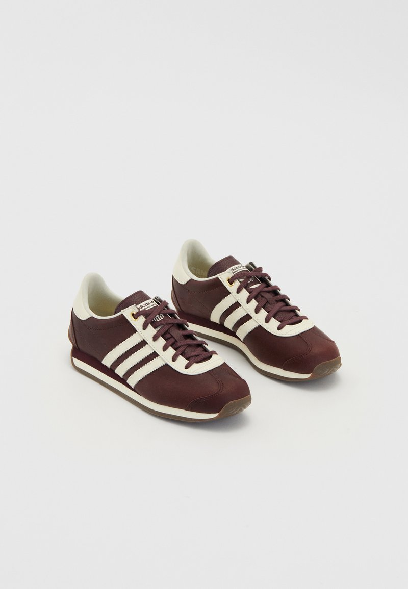 Sötét bordó sneaker fehér csíkokkal, fűzős kialakítással, párnázott bokarész és textúrált gumi talppal. Simább anyag, ellentétes színű díszítésekkel.