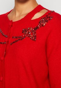 Cardigan rouge avec un col rond, une fermeture à boutons, et des accents décoratifs en sequins formant un nœud sur le devant et la zone de l'épaule.