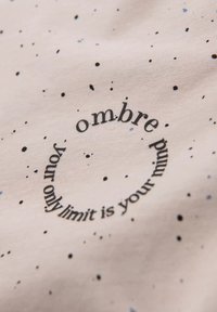 Lichtroze stof met verspreide zwarte en blauwe spikkels. Bevat een cirkelvormige zwarte tekst: "je enige limiet is je geest ombre."