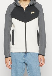 Mikina Nike na zip s barevným blokovým designem v šedé, černé a bílé barvě, s bočními kapsami a kapucí. Zlaté logo na hrudi.