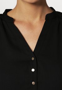 Czarna bluzka z kołnierzem typu stójka i pięcioma metalowymi zatrzaskami z przodu. Tkanina wydaje się gładka i lekka.