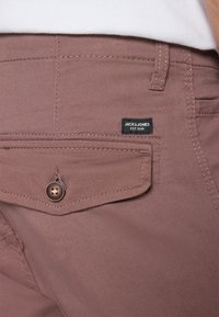 Bordeauxkleurige broek met een voorzak met een bruine knoop, gestikt detail en een zwart label met "JACK & JONES EST. 1990."
