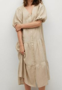Läggdags-beige linneklänning i knälängd med korta puffärmar, djup V-ringning och knappfront. Har en tiered kjoldesign och mjuk textur.