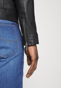 Black leather jacket with a zippered cuff detail, paired with blue denim jeans. The hand rests near the jacket's zipper accent.