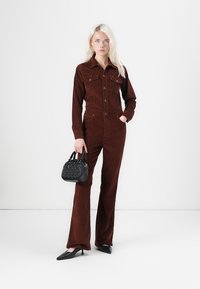 Bruine corduroy jumpsuit met lange mouwen, voorkruiszakken en een getailleerde taille, gecombineerd met een zwarte geplooide handtas en zwarte puntige schoenen.
