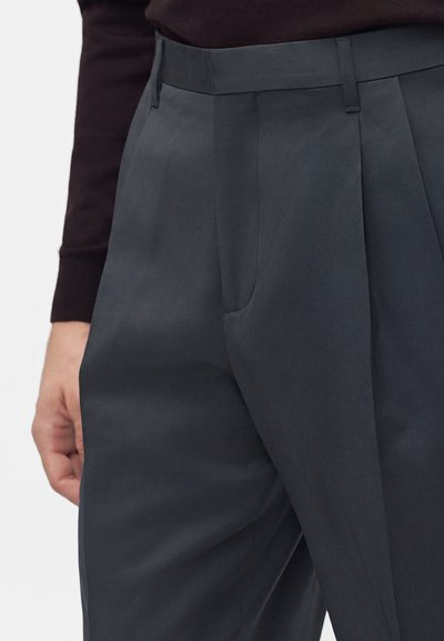Gros plan sur un pantalon gris foncé cintré avec des plis à l’avant et des passants de ceinture, porté avec un haut noir à manches longues.