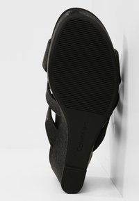 Semelle de sandale à talon noir avec grip texturé et logo Calvin Klein sur fond blanc, chaussure positionnée verticalement contre un mur.