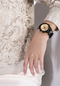 Schwarze Gummiband-Uhr mit einem goldfarbenen runden Ziffernblatt, großen Ziffern und Krone, die an einer Hand vor einem Spitzenstoff-Hintergrund getragen wird.