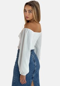 Blouse blanche à épaules dénudées avec de longues manches bouffantes, détail smocké à la taille, associée à une jupe en denim taille haute avec fente à l'avant.
