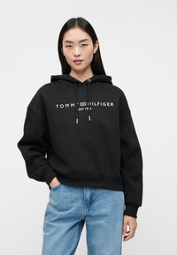 Juodas su kapuce megztinis su laisvu siluetu, puošiamas baltu „TOMMY HILFIGER“ logotipu krūtinėje ir su raištelių detalėmis.