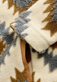 Fleecejacke mit cremefarbenem Grundton und strukturierten braunen und grauen Sternmustern. Mit einem glatten braunen Akzent an der Innenseite.