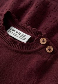 Le pull en tricot bordeaux présente un motif texturé, un col côtelé et deux boutons en bois à l'épaule. L'étiquette indique la taille et l'origine.