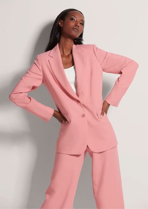 Vrouw poseert met handen in de zij, draagt een lichtroze blazer en bijpassende wijde broek boven een wit topje.