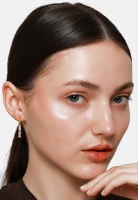 Gouden hoepel oorbellen met heldere kristalaccenten, die een verticale ontwerpen laten zien. Het model heeft een egale huid en een neutrale lipkleur.