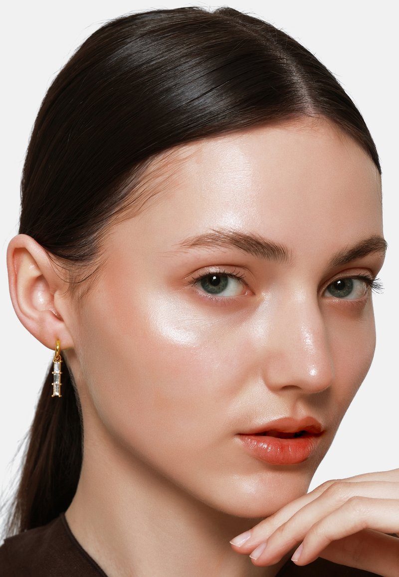 Gouden hoepel oorbellen met heldere kristalaccenten, die een verticale ontwerpen laten zien. Het model heeft een egale huid en een neutrale lipkleur.