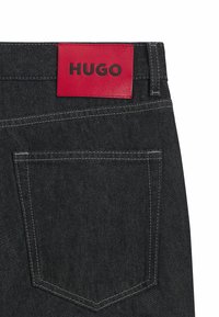 Jeans in denim nero con una toppa in pelle rossa etichettata "HUGO" in vita. Presentano dettagli di cucitura e una tasca posteriore.