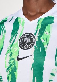 Baltas sportinis marškinėliai su žaliais abstrakčiais dryžiais. Savyje turi apvalų logotipą su ereliu, "Nigerijos futbolo federacija" ir "1945" juoda spalva. Juoda Nike ženklas.