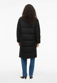 Ilgi melnā pūkainā mēteli ar horizontālu quilted dizainu. Iezīmē garas piedurknes un taisnu griezumu, apvienojot to ar ziliem taisnas kājas džinsiem un melnām augstpapēžu kurpēm.
