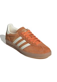 Brune ruskinds sneakers med hvide striber og orange snørebånd. Har en rund tå, polstret krave og gummisål med præget logo.