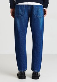 Jean en denim bleu foncé avec une coupe décontractée, présentant deux poches arrière et un léger délavage, associés à des chaussures noires et un pull texturé.
