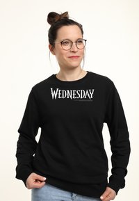 Donna con occhiali e capelli raccolti in uno chignon, indossa una felpa nera con la scritta "WEDNESDAY", tenendo l'orlo, mentre guarda leggermente verso destra.