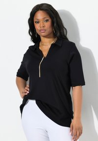 Maglietta polo nera con colletto con zip, maniche corte e orlo leggermente curvo. Il modello indossa collane d'oro a strati e pantaloni bianchi.