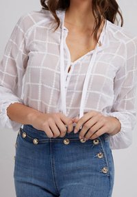 Femme portant un chemisier blanc transparent à texture avec des boutons et un jean bleu taille haute avec des boutons dorés à la ceinture et sur le côté.
