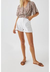 Femme portant un haut cache-cœur à fleurs avec des manches volantées, un short blanc taille haute retroussé et des sandales plates tressées marron sur fond blanc.