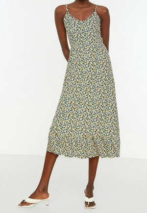 Mujer de pie vistiendo un vestido floral sin mangas de largo midi con pequeñas flores blancas y amarillas y sandalias blancas de tacón.