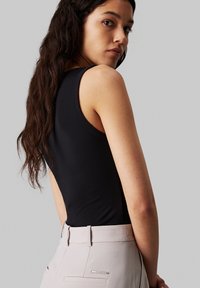 Musta ilmanhiha bodysuit, jossa on alhainen selkäosa, yhdistettynä beigeihin istuviin housuihin, joissa on huomaamaton takatasku ja hopeiset yksityiskohdat.